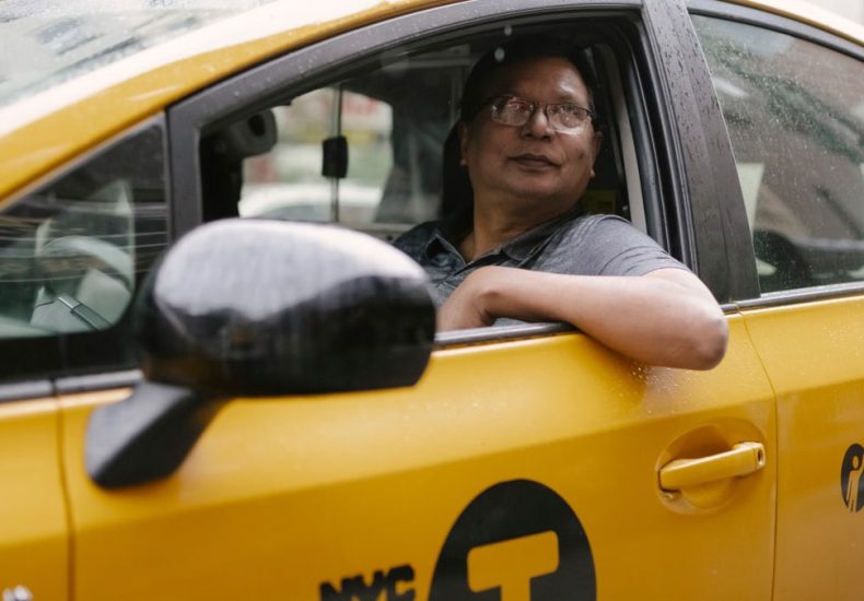 Serious taxi driver in glasses sitting in yellow automobile and looking up on street in city in daytime